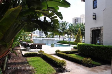 A pale stone walkway runs between lawn and clipped hedges beside a pool terrace and a planted house wall.