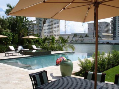 Pale stone paving covers a poolside dining terrace beneath a large umbrella, with palms and waterside buildings beyond.