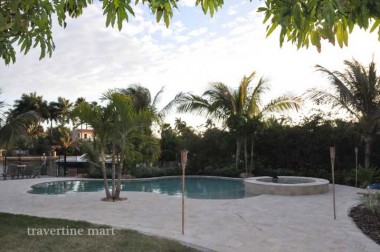 Light mixed-size paving surrounds a freeform pool, palm trees and a raised round spa in a landscaped garden.