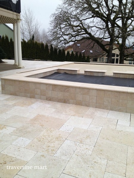 Pale mixed-size pavers cover a terrace beside a square raised stone-faced basin and a leafless garden tree.