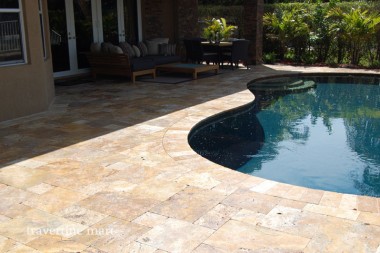 Golden mixed-size pavers follow a curved pool edge beside covered cushioned seating and dense planting.