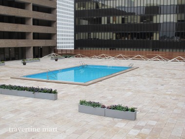 Pale rectangular pavers surround a square pool between multi-level buildings, with long flower planters along the deck.
