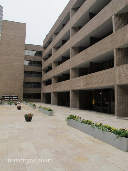 Light rectangular pavers cover a broad courtyard terrace beside a multi-level building and long planters.