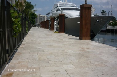 Pale mixed-size pavers cover a narrow waterside walkway between a dark railing and a docked white boat.