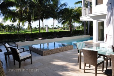 Large pale square pavers form a shaded seating terrace beside a raised mosaic-faced spa, palms and a white house.