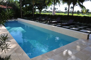 Pale square pavers edge a long rectangular pool with a raised back wall and loungers set between lawn strips beneath palms.