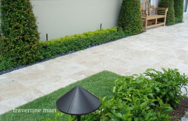 A pale paved garden walkway runs beside a white wall, clipped hedges and a small timber garden gate.