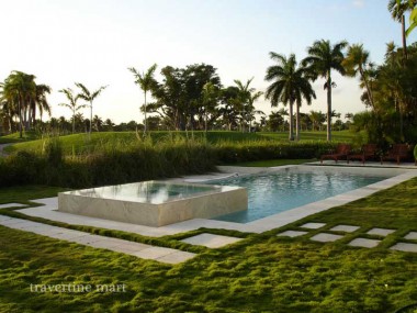 A rectangular pool and raised square basin are bordered by pale stone edging and stepping slabs set within lawn.
