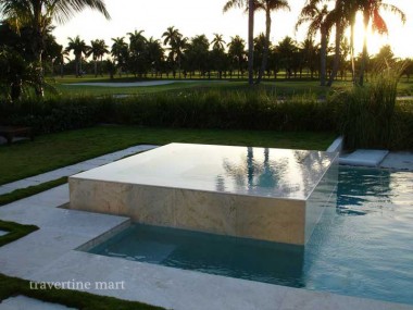 A pale stone-clad square basin projects into a rectangular pool, reflecting palms and evening light beside lawn.