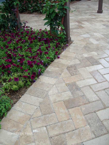 Small cream and brown rectangular pavers meet a curved planting bed filled with purple flowers and green foliage.