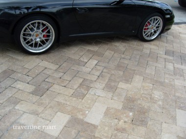 Cream and brown rectangular pavers are laid beneath a dark car, showing the staggered joints and varied stone tones.