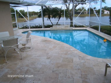 Pale mixed-size pavers surround a pool inside a screened enclosure with white chairs and open water beyond.