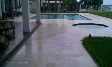 Pale mixed-size stone paving links a rectangular pool, an adjoining round spa and a covered patio with white columns.