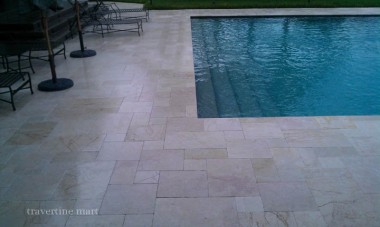 Pale mixed-size paving follows the straight edge of a rectangular pool, with outdoor dining furniture along the terrace.