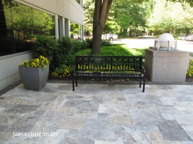 Gray and cream mixed-size pavers cover a terrace with a black garden bench, large planters and a tree-lined garden.