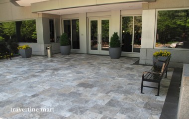 Gray mixed-size paving extends across a covered terrace outside glass doors, with large planters and a wooden chair.