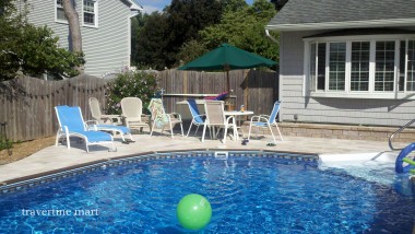 Light stone paving follows a curved blue pool beside white patio chairs, a green umbrella and a timber fence.