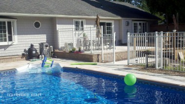 Pale mixed-size paving surrounds a blue pool beside an elevated patio enclosed by light metal railings.