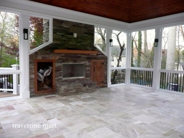 Mixed-tone pale tiles cover a screened porch with a stone fireplace, a wood ceiling and views of trees outside.