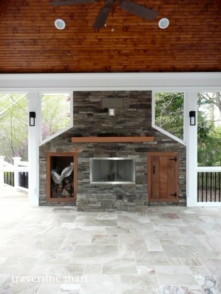 Light mottled floor tiles surround a stone fireplace beneath a wood ceiling in a screened porch with white framing.