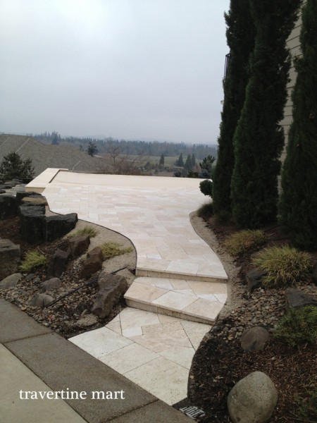 A pale paved path curves down a short set of garden steps between rock landscaping and tall evergreen trees.