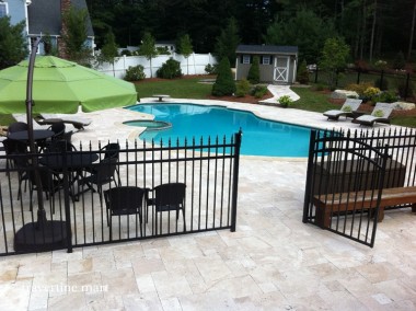 Pale mixed-size paving leads through an open black metal gate to a pool deck with a green umbrella and chaise lounges.