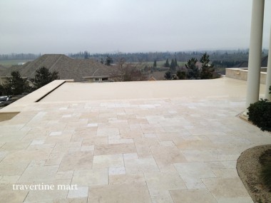 Cream mixed-size paving spreads across an open raised terrace with distant rooftops and a column along the house.