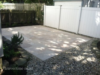 Pale mixed-size pavers form a shaded garden patio bordered by a white fence, gray pebbles and low plants.