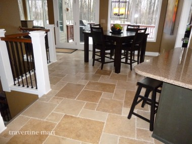 Brown mixed-size floor tiles continue beneath a dining table beside kitchen counters and a stair railing.