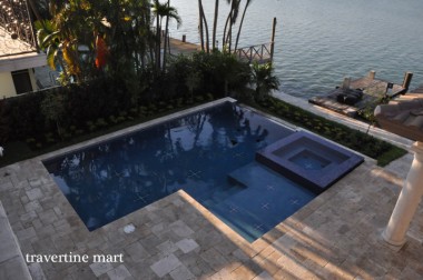 An overhead view shows a dark-blue pool, raised square spa and mixed-size stone deck beside palms and a waterside dock.