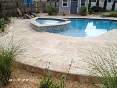 A raised round spa and a broad pale stone terrace sit in a fenced backyard with planting beds and a small shed.