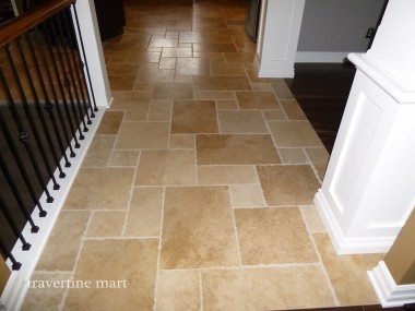 Warm brown mixed-size tiles cover a hallway between white walls and a dark stair railing.