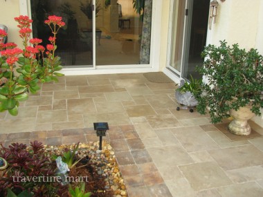 Brown mixed-size tiles cover a patio outside sliding doors, bordered by red flowers, potted plants and decorative pebbles.