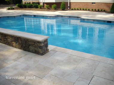 Pale stone paving borders a pool and a short stacked-stone wall topped by long light-colored caps.