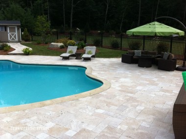 Pale stone paving surrounds a curved blue pool and dark outdoor seating shaded by a green umbrella.