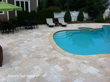 Cream and tan mixed-size pavers follow a curved blue pool, with chaise lounges and a green umbrella nearby.