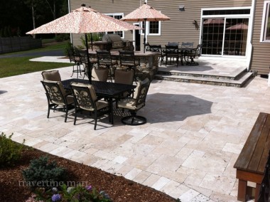 Pale mixed-size pavers cover a dining patio with metal tables, chairs and patterned umbrellas beside a house step.