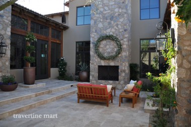 Light rectangular pavers cover a courtyard with a tall stone fireplace, broad house steps, potted plants and wooden seating.