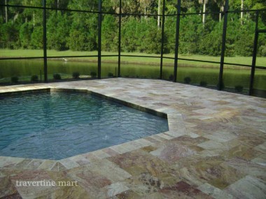 Cream, gold and reddish mixed-size pavers border an angular pool inside a screened enclosure overlooking a lawn.