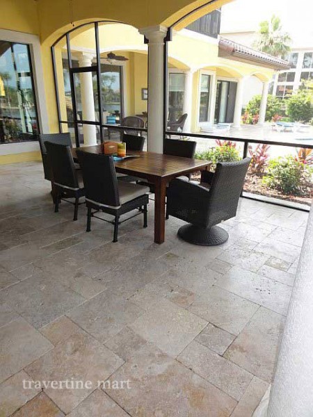 Light mixed-size pavers cover a shaded dining patio with dark chairs, a wood table and an arched house opening.