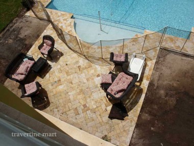 An overhead view shows small golden pavers beneath cushioned outdoor chairs beside broad shallow pool steps and a mesh barrier.