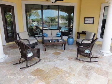 Pale square pavers cover a columned patio with woven chairs, a low table and blue decorative planters.