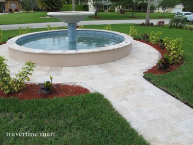 A circular tiered fountain stands within pale paving that connects to a narrow garden path between lawn and flower beds.