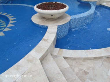 Light stone coping curves around blue mosaic-lined water with a round planter at the junction of the basins.