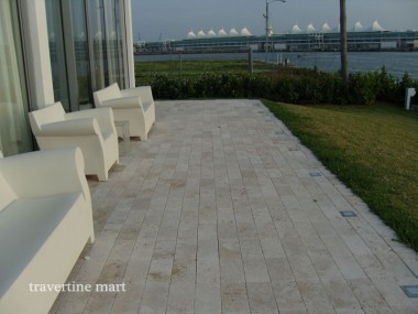 Small rectangular pale pavers cover a waterside patio beside white lounge chairs, house glazing and a strip of lawn.