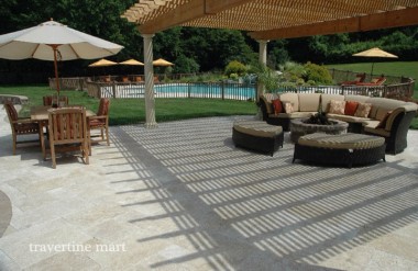 Pale rectangular pavers cover a pergola terrace with woven seating and a timber dining table beside a fenced pool.