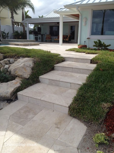 Pale stone steps climb between lawn and rocks to a covered patio with white posts and dark outdoor seating.
