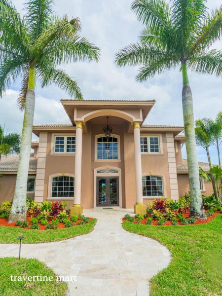 A pale stone walkway curves across the lawn toward a tall arched entry framed by palms and flowering shrubs.