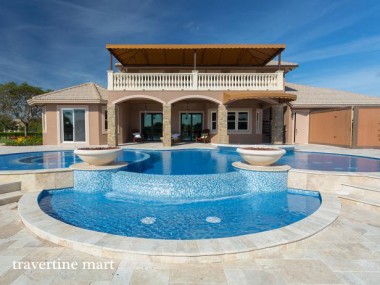 Pale paving wraps a blue pool and a curved raised water feature in front of a house with arches and an upper terrace.