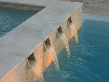 Wide pale coping tops a raised pool wall with a row of narrow spillways pouring into blue water.